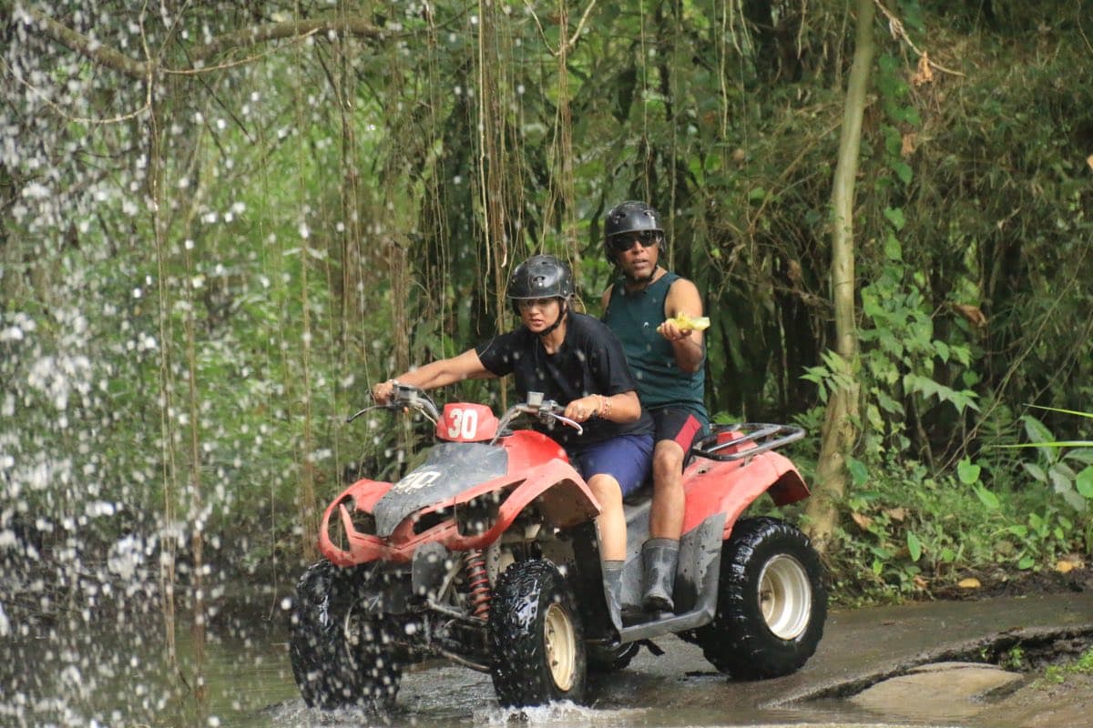 Two riders on an ATV at a roadside jungle waterfall