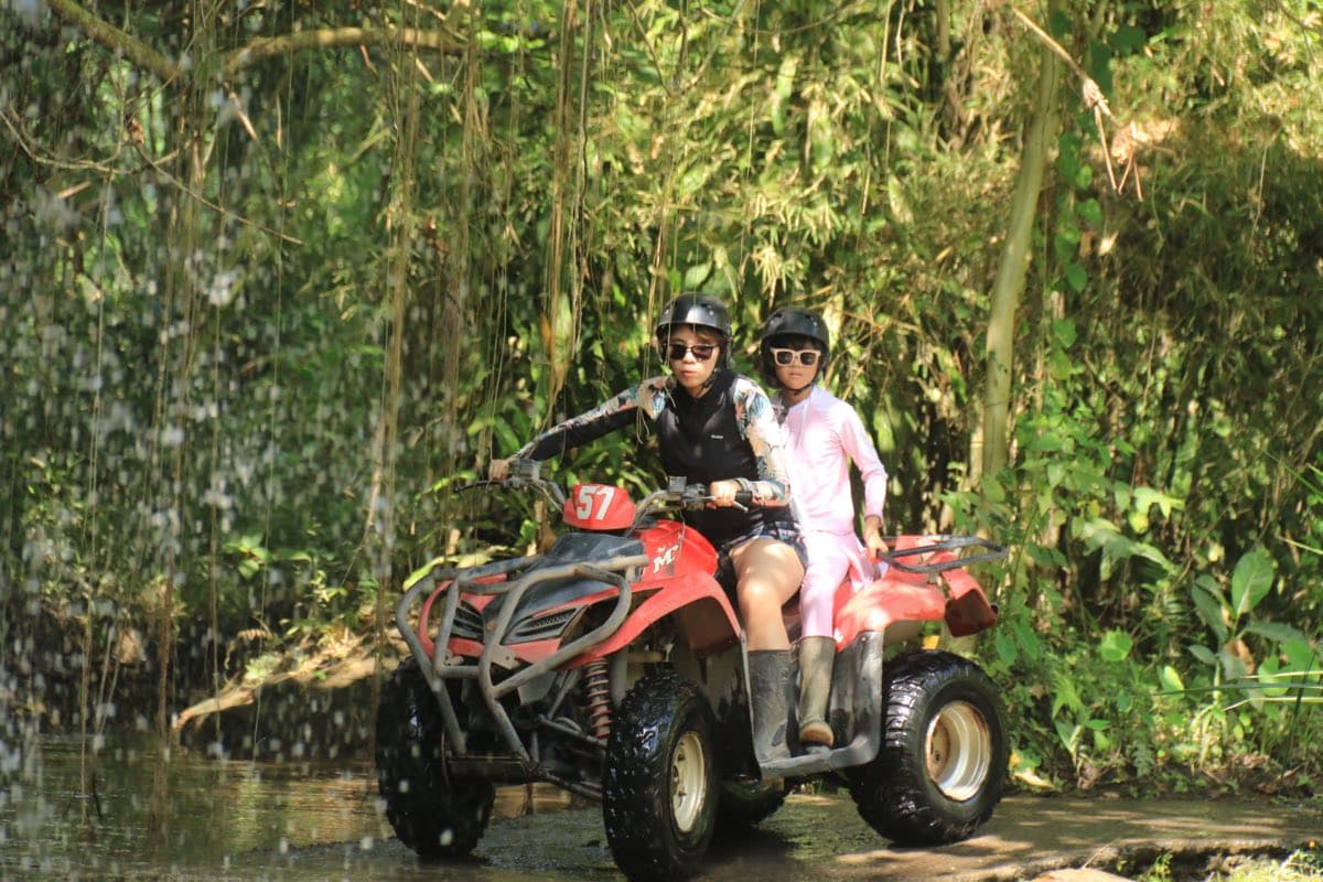 Two riders on a red ATV beside a jungle waterfall