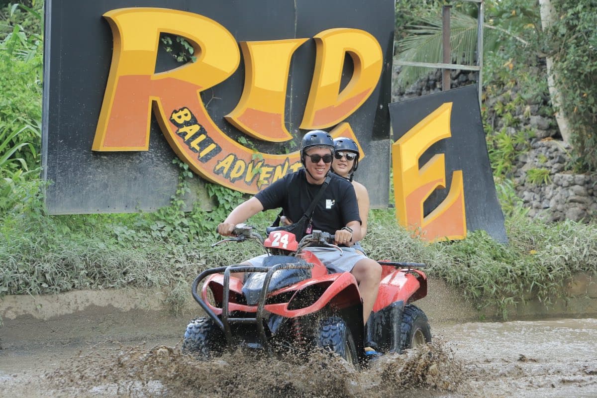 Two riders on an ATV splashing through a muddy pool