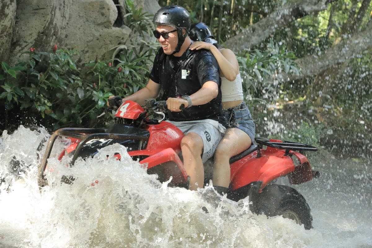 Two riders speeding an ATV through a splashing river