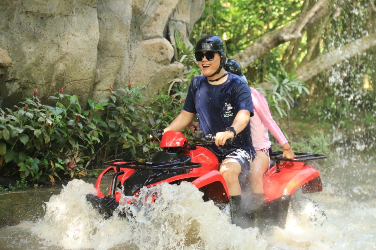 Two riders splashing a red ATV through a jungle waterfall
