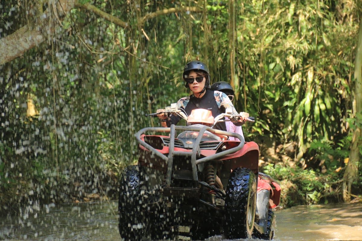 Two riders pausing on a red ATV beneath a jungle waterfall